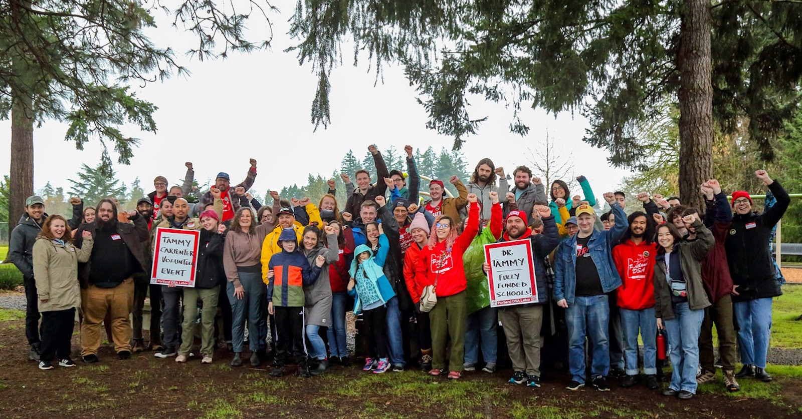 A group of approximately 40 people are gathered outside and smiling with their left fists raised. Two people hold DSA-branded signs that read "Volunteer Event for Tammy Carpenter" and "Dr. Tammy. Fully Funded Schools" Many participants are wearing bright red Portland DSA merch.